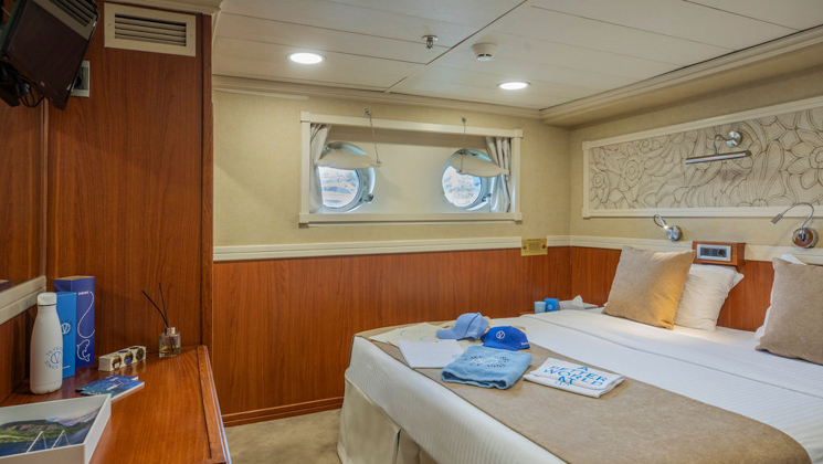 Category B cabin with double bed in beige, wood bedside tables & desk, silver floral accent wall & 2 portholes on Galileo ship.