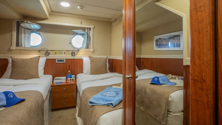 Category C cabin with 2 twin beds in beige, wood bedside table & 2 portholes on Galileo cruise ship.