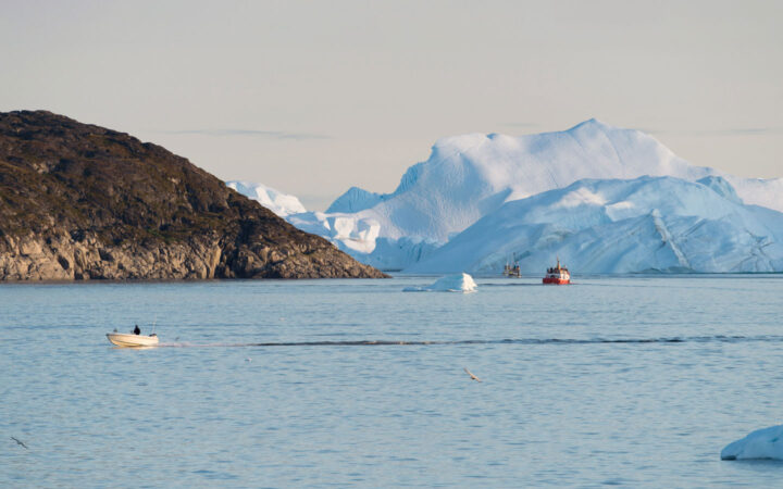 White day boat cruises calm water in morning light as red & white fishing boats troll beside large icebergs & rocky cliffs.