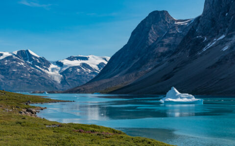 Iceberg sits in blue water on a windy day beside green shores & dark, rocky mountains partly covered in snow, seen in Greenland.