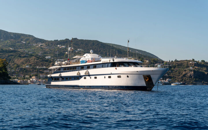 Harmony G white & black mega yacht with 3 guest decks sits at anchor in Mediterranean port on a sunny day.