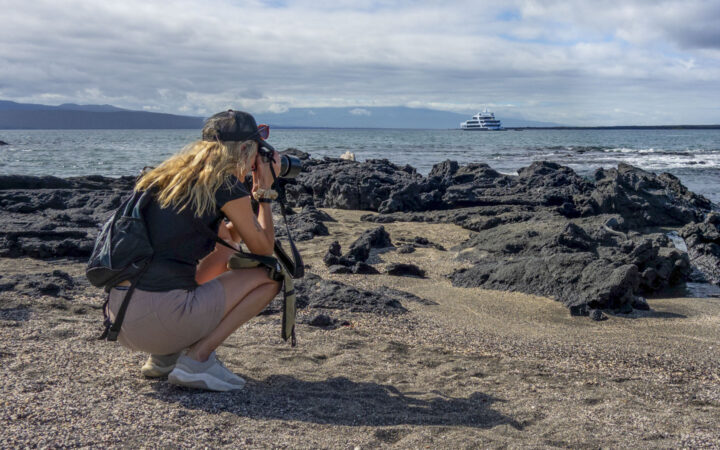 A female traveler squats on the sand taking a photo with Hermes catamaran in the water behind her