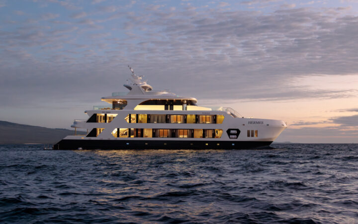 Hermes yacht in Galapagos at dusk with 4 white upper decks, 2 interior lit & 2 al fresco, and dark blue waterline deck.