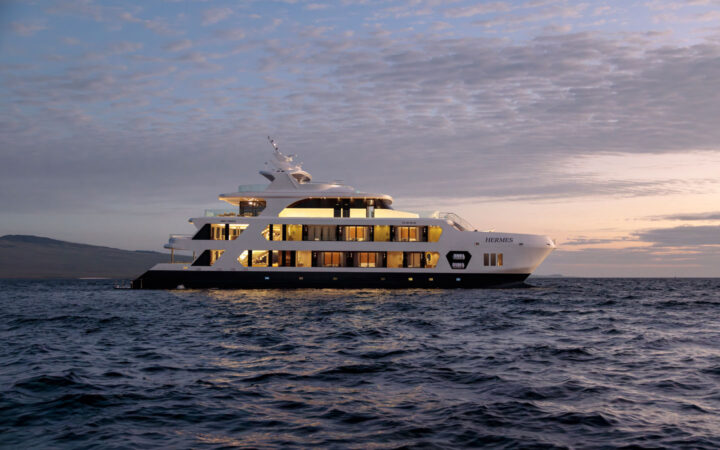 Hermes yacht in Galapagos at dusk with 4 white upper decks, 2 interior lit & 2 al fresco, and dark blue waterline deck.
