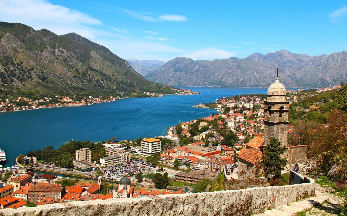 View from beige stone walkway down to a seaside town in the Mediterranean, with green hills, church steeple & terra cotta roofs.