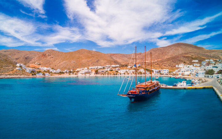 Aerial view of small motorsailor with blue hull & teak decks docked in teal sea at a small island town in Greece's Cyclades.