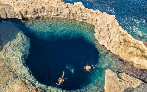 Aerial view of 2 Malta cruise guests swimming in a dark blue caldera beside open ocean during the La Dolce Vita cruise.
