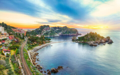 Aerial view of Taormina, Sicily at sunset with rocky, green islands & rail line along idyllic bays, seen on a cruise to Malta.