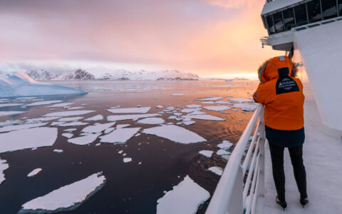 Woman in orange parka with fur-lined hood stands on deck of white ship photographing sea ice at sunset in the Bellingshausen Sea.