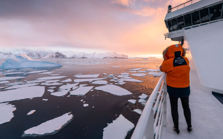 Woman in orange parka with fur-lined hood stands on deck of white ship photographing sea ice at sunset in the Bellingshausen Sea.