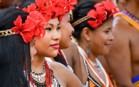 Indigenous women in Panama with brightly colored beads, woven headbands with red flowers & colorful clothes stand in a line.