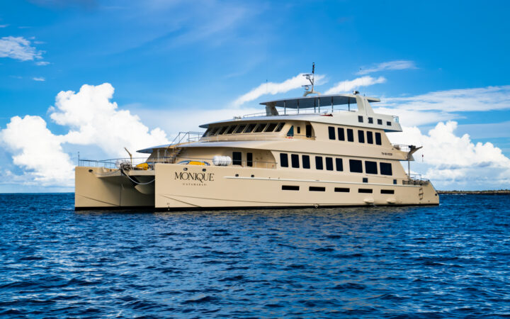 Monique luxury catamaran floating on on calm blue waters in Galapagos with cream-colored hull & multiple decks.