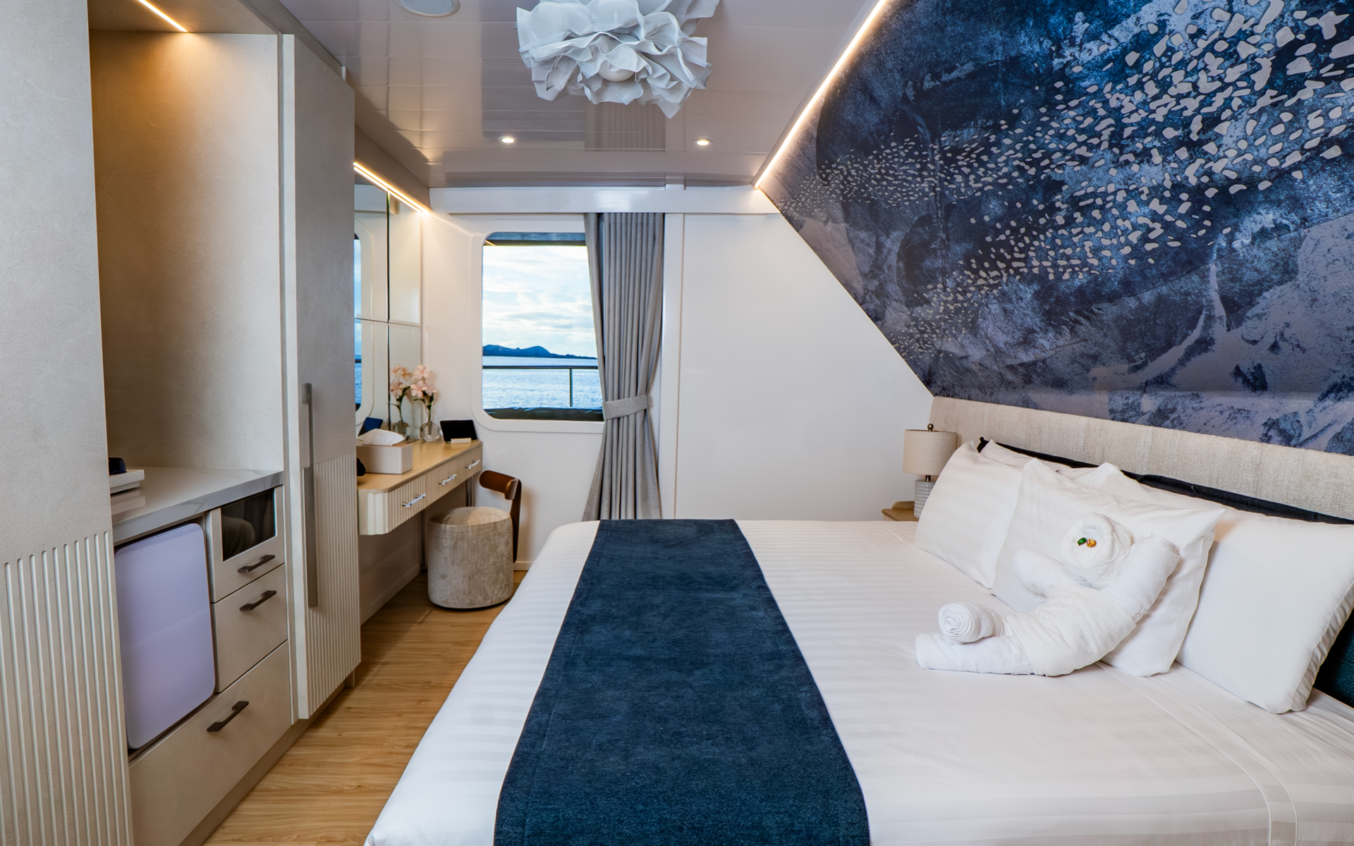 King bed next to closet, desk & a large window looking out to the Galapagos waters in Golden Single Stateroom aboard Monique yacht.