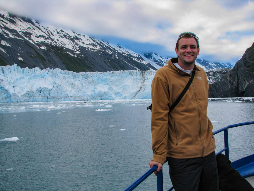 Man in tan jacket stands beside blue railing on bow of M/V Discovery ship in front of a glacier in Alaska.