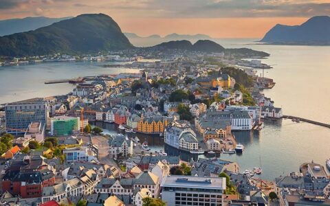 Aerial view of water-surrounded small town of various colorful buildings seen at sunset, surrounded by mountains.