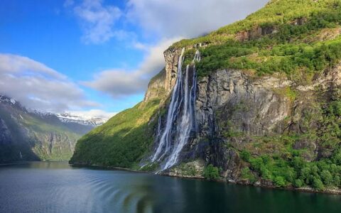 Large waterfall with 2 main falls spews over cliff face into glassy water by green hillsides on a Norwegian fjords cruise.