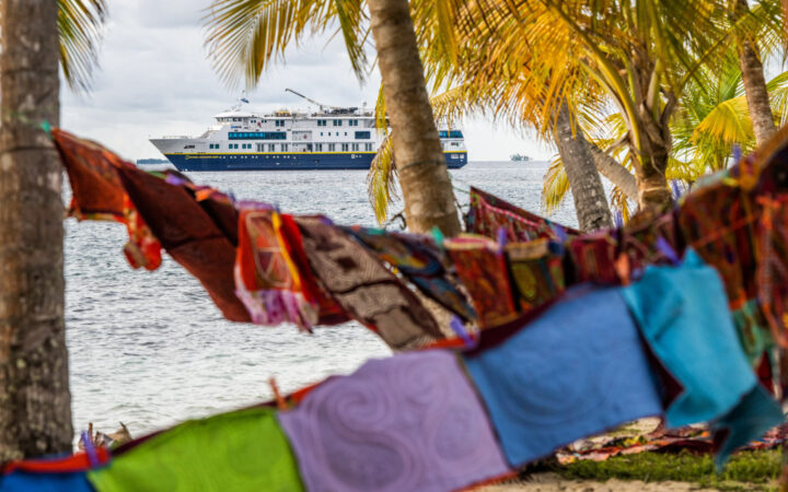 Colorful, handwoven squares hung on lines between palm trees in foreground & small blue, gold & white Nat Geo ship in background.