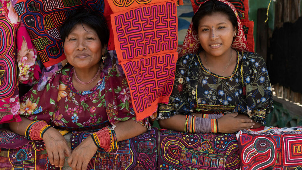 2 women sit & smile among brightly colored, woven textiles, seen on a Panama & Colombia small ship cruise.