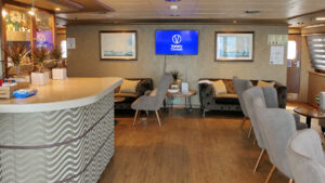 Bar of Panorama Mediterranean sailboat with silver & gold waves, wood floors, gray velvet chairs, TV & framed art.