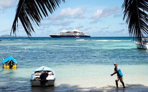 A man walks toward a dinghy on a white-sand beach with palm tree fronds above & a luxury ship in the distance during the Pearls of the Caribbean luxury cruise.