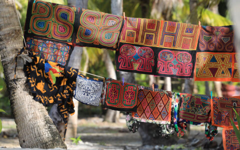 Multicolored, bright, woven textile squares hang from rope between palm trees over white-sand beach in Panama.