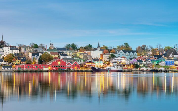 Hillside harbor town at dawn with docked fishing boat, colorful homes, church steeples, fall foliage & glassy water.