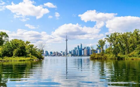 Toronto's CN Tower rises above a glassy river, green forested riverbanks & city high rises, seen on small ship great lakes cruises.