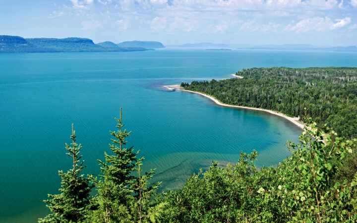 Aerial view looking down on Lake Superior on a calm day with its turquoise water, white-sand shoreline & bright green forest.