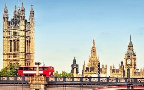Red double-decker bus drives across a bridge in London with Big Ben & Victoria Tower in the distance on a sunny day.