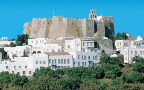 A view of Patmos and its medieval town of Chora behind more modern white buildings and green trees.