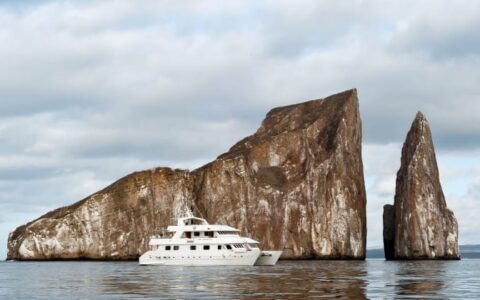 The seaman journey cruise, a luxury yacht, motors across a calm ocean in front of rocks jutting out of the water at the Galapagos islands