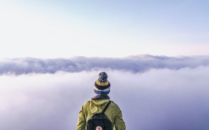 The back of a person with a beanie hat, green jacket and backpack standing on top of a mountain overlooking a dramatic cloud covered landscape below.