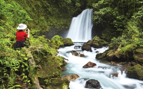 A female traveler in a red shirt and white wide brimmed hat kneels in lush green forest photographing a waterfall in the jungle of Costa Rica.