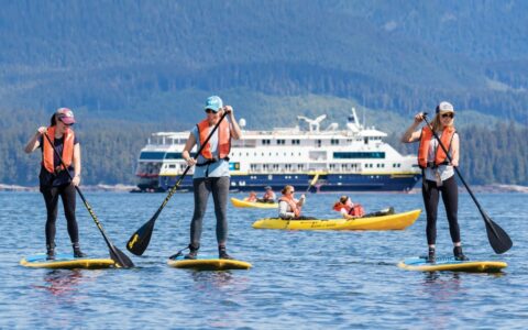 Three women stand-up paddleboarding with a National Geographic expedition ship in the background, on a sunny day.