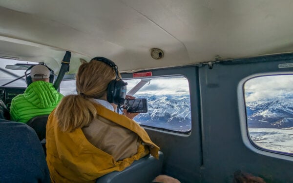 Women looking out of small aircraft with yellow jacket and headset on and her phone capturing the snowy mountains