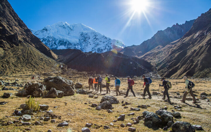 line of adventure travelers walking along a sunny path with a large snowy mountain in the distance on the salkantay trek to machu picchu peru trip