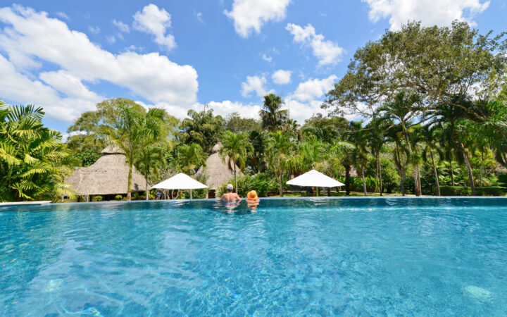 Two travelers stand at the edge of an infinity pool with blue skies above & lush jungle ahead during the Chaa Creek Inland Expedition.