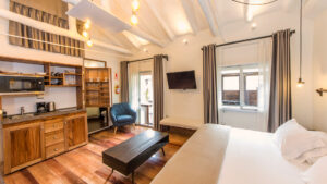 Superior Deluxe room at XO Art Hotel Cusco with king bed in white, coffee table, kitchenette, balcony, chair & hanging lights.