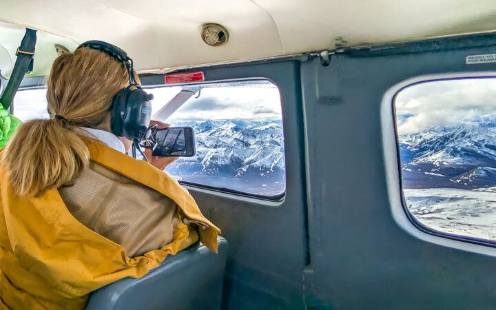 Women looking out of small aircraft with yellow jacket and headset on and her phone capturing the snowy mountains