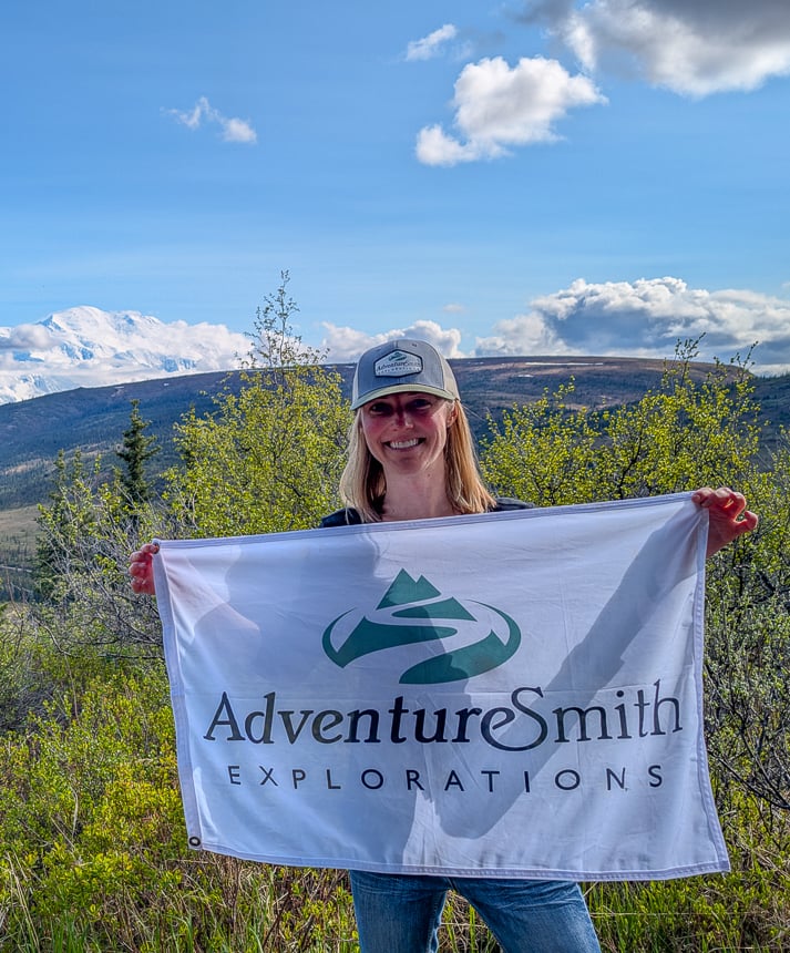 Picture of AdventureSmith Exploration Lauren in Alaska with AdventureSmith flag and hat on, on a sunny day in front of Denali 