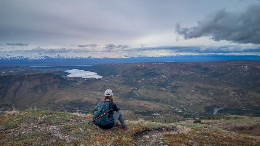 Hiker sitting with her backpack on looking out over lake wonder and camp denali on a cloudy day 