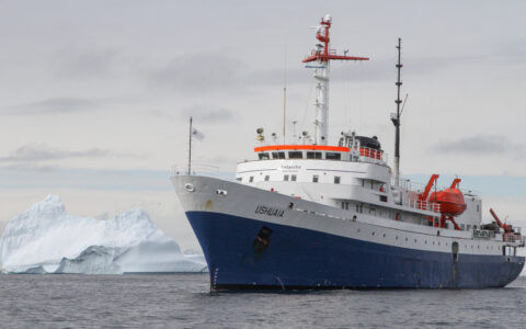 Ushuaia small ship in blue & white sits beside a large iceberg on the Classic South Georgia small ship cruise in Antarctica.