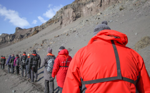 Line of Classic South Georgia cruise guests in colorful jackets walk a trail over gray dirt under blue skies.