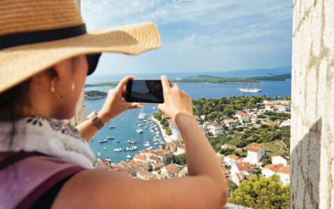 Woman in woven sunhat holds phone to photograph Hvar, Croatia with beige stone buildings, terracotta roofs & green trees.