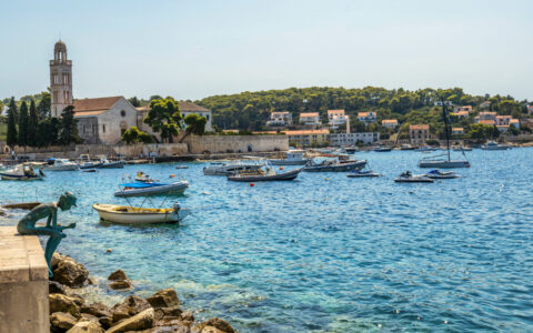 Weathered stone statue of a person sits overlooking teal sea & small fishing boats on moorings beside sunny, seaside town.