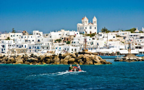 Red Zodiac with Greece & Turkey cruise guests makes its way towards small harbor of whitewashed hillside town in Greece.