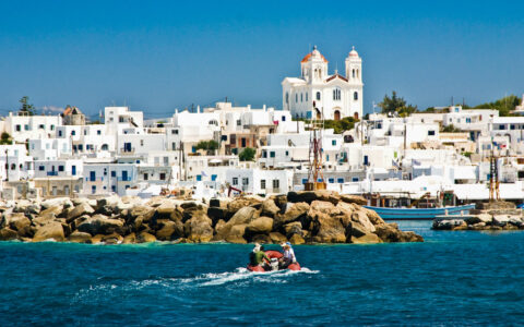 Red Zodiac with Greece & Turkey cruise guests makes its way towards small harbor of whitewashed hillside town in Greece.