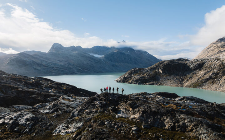 Aerial view of small group of hikers standing atop a rocky mountain overlooking a fjord with teal water in East Greenland.