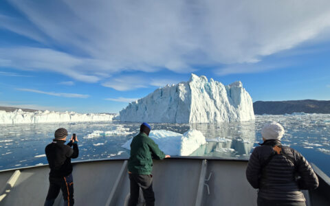 3 East Greenland Sermilik fjord cruise guests stand on bow of small ship in warm clothes, photographing large icebergs.