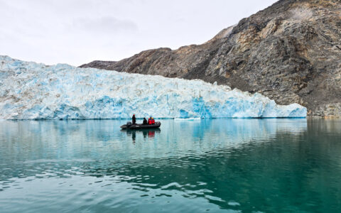 Zodiac boat with East Greenland cruise guests idles in glassy teal water beside large, chunky blue glacier & dark cliffs.